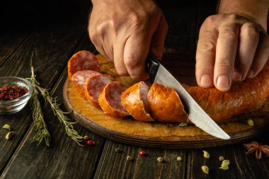 Ev mutfağındaki ahşap kesme tahtasındaki sosisi bir aşçının elleriyle dilimlemek. Fast food pişirme fikri.