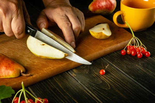 The cook cuts a pear with a knife on a cutting board to prepare compote or fruit juice. The concept of a pears diet based on a set of vitamins. Work environment on the kitchen table.