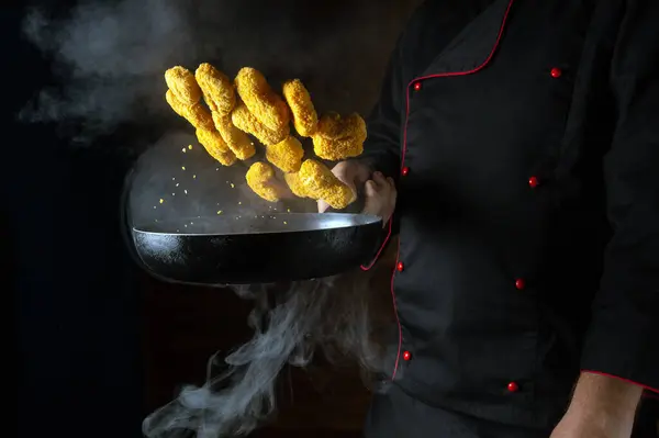 The hand of a professional chef throws nagits in a hot frying pan with steam on a black background. The concept of cooking in the hotel. Free ad space.