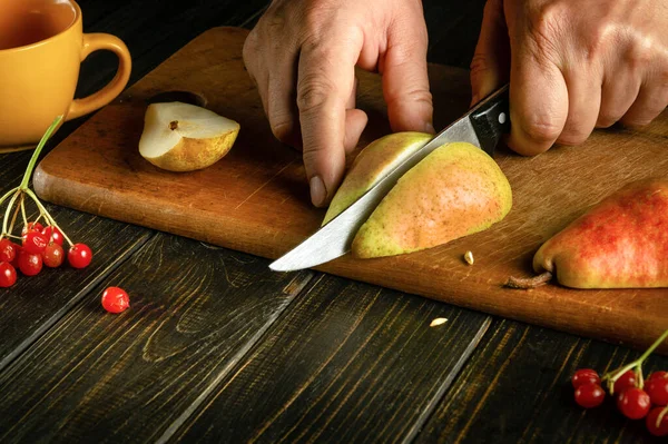 The chef hands use a knife to cut a ripe pear on a cutting board to prepare compote or fruit juice. Pear diet or fruit dessert