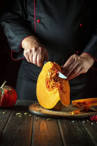 A chef cuts an orange pumpkin into slices with a knife on a wooden cutting board before preparing porridge for breakfast. Autumn food concept in cozy dark kitchen.