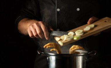 Şef kaynayan tavuk bacağına soğan ekliyor. Restoran mutfağında yağlı et suyu pişirme kavramı ve siyah arka planda reklam alanı..
