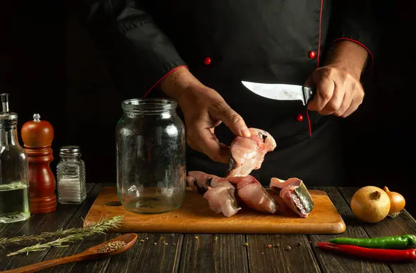The chef cuts raw fish into steaks before preparing herring in a jar. Working environment on the kitchen table with aromatic spices and pepper.