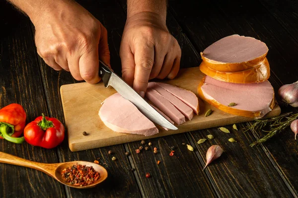 Slicing parboiled sausage with a knife in the hands of a chef for preparing a delicious lunch on the kitchen table. Fast food concept.