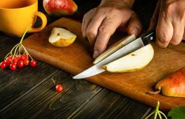 Kesme tahtasındaki bir armudu kesmek için bıçak kullanan bir adamın ellerini komposto veya meyve suyu hazırlamak için yakın plan çek. Armut diyeti kavramı bir vitamin kompleksine dayanır. Siyah mutfak masasında çalışma ortamı.