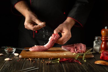 Şef, barbekü yemeği için bifteğe tuz serpiyor. Mutfak masasında biberiye ve sarımsaklı çalışma ortamı.