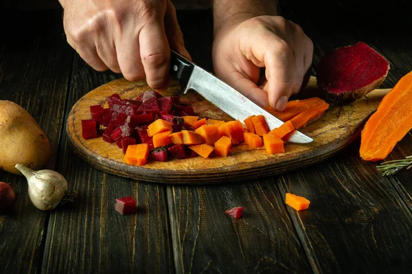 A cook uses a knife to cut boiled carrots to prepare a vegetable dish on the kitchen table. Venaigret cooking concept on dark background.