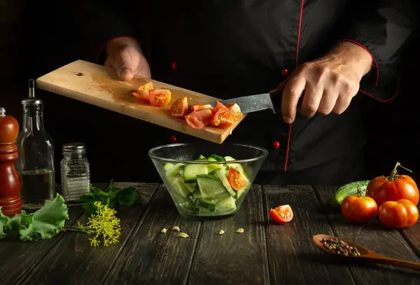 A professional chef prepares a salad from fresh vegetables in the kitchen. The concept of preparing a delicious salad with tomatoes and cucumbers. Place for advertising or recipe on a dark background.
