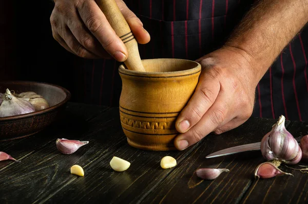 The cook crushes the garlic on the kitchen table before adding it to the food. Close-up of a man hands chopping garlic in a wooden mortar. Peasant food concept on dark background.