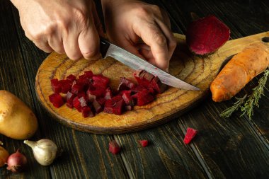 Tahta bir kesme tahtasının üzerinde kaynamış kırmızı pancarları bıçakla kesen bir adamın ellerinin yakın çekimi. Tatilden önce evde venegret hazırlama konsepti.