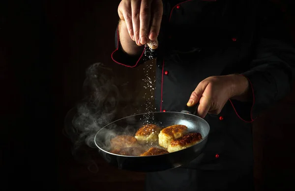 The cook salts hot pancakes in a frying pan while frying. Concept of cooking oladki in a restaurant kitchen. Black space for menu or recipe.