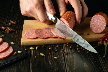 Aşçı ahşap bir kesme tahtasının üzerinde bir bıçakla et sosisi keser. Kahvaltı için lezzetli sandviçleri evdeki mutfak masasında hazırlıyorum..
