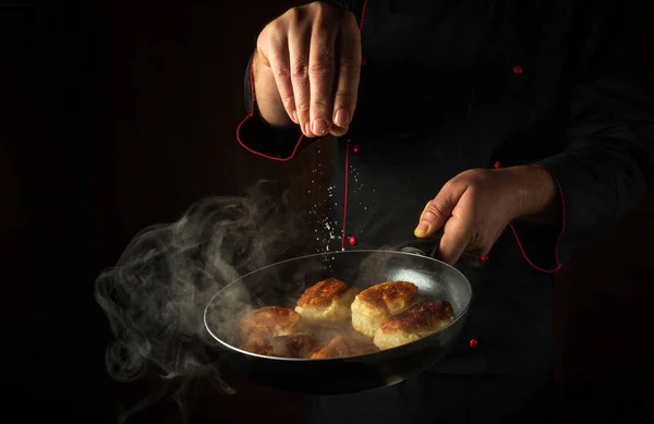The cook adds salt to oladki in a frying pan. The concept of preparing a delicious Ukrainian national dish or bliny in the kitchen. Black space for advertising.