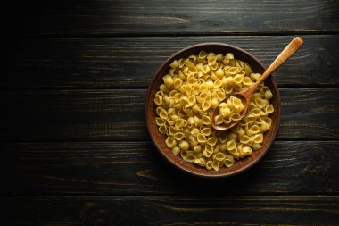 Pişirmeden önce bir tabak çiğ kabuklu makarna ve antika bir masada bir kaşık. Koyu arkaplanda tarif veya menü için yer.
