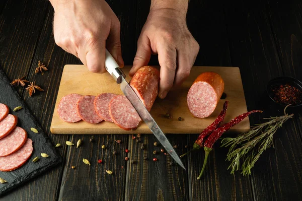 Cooking a hearty breakfast with smoked sausage by the hands of a cook on the kitchen table. Cutting sausage with a knife for a serving board.