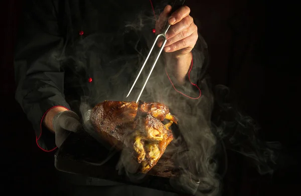 A professional chef prepares chicken in a restaurant kitchen. A hot baking sheet with a baked bird carcass and a fork in the hands of a cook.