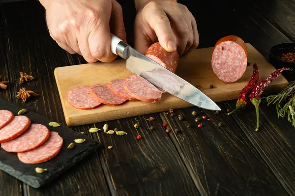 Şefin elleri bir sandviç için füme sosisi ince dilimlere ayırmak için bıçak kullanır. Karanlık masada fast food pişirme konsepti.