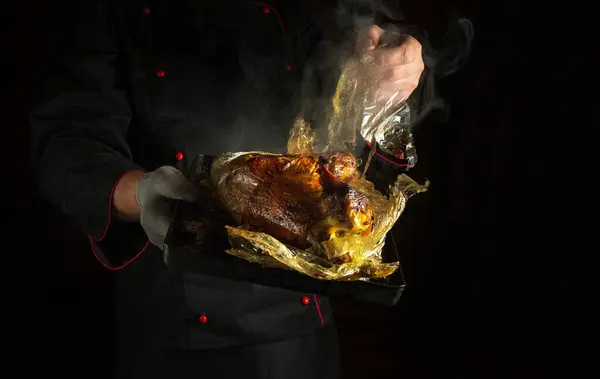 The chef manually removes the plastic bag from the chicken after baking on a baking sheet in the oven. Concept of grilling poultry in a restaurant or hotel kitchen. Dark space for recipe or menu.