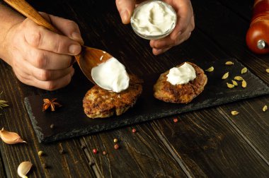 Bir aşçının eli, servis tahtasındaki et pirzolalarına kaşıkla ekşi krema ekliyor. Lezzetli et ziyafeti konsepti.