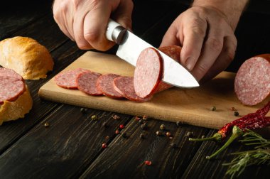Şef ahşap bir kesme tahtasıyla et sosisi keser. Bir sokak restoranının mutfak masasında kahvaltı için lezzetli sandviçler hazırlıyoruz..