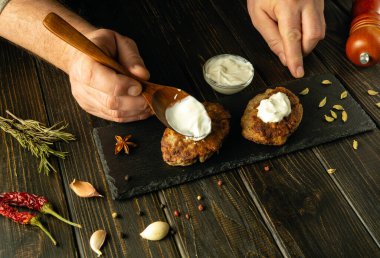 Erkek eller, ayıklama tahtasındaki kızarmış pirzolalara bir kaşıkla ekşi krema ekler. Lokantada lezzetli yemekler hazırlama kavramı.