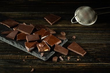 Siyah mutfak masasında kırılmış siyah çikolata parçaları ve çikolata parçacıkları için mutfak süzgeci. Tatlı pişirme konsepti