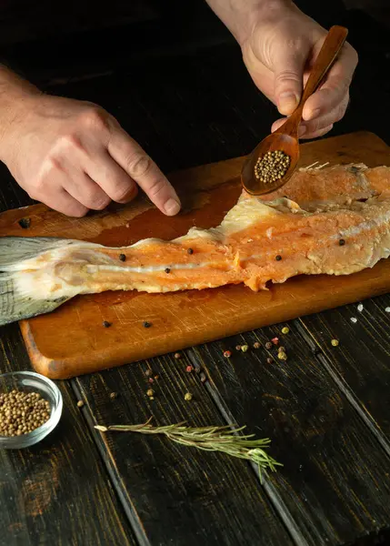 The cook hands add coriander with a spoon to a raw fish carcass on a cutting board. Concept of preparing fish dish in saloon kitchen.