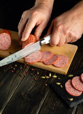 Şef ahşap bir mutfak kesme tahtasına bıçakla salam et sosisi keser. Hızlı bir aperatif için basit bir masa ayarlama kavramı.