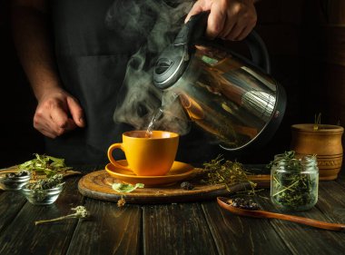 Bir adam mutfak masasında sağlıklı çay yapmak için kupaya kaynar su ekler. Geleneksel tıp konsepti ya da tıbbi çayın tıbbi bitkilerle hazırlanması.