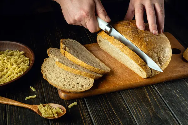 Aşçı elleri akşam yemeği için mutfak tahtasına bıçak ile taze ekmek kesti. Sağlıklı yiyecekler ve geleneksel fırın ürünleri konsepti. Boşluğu kopyala.