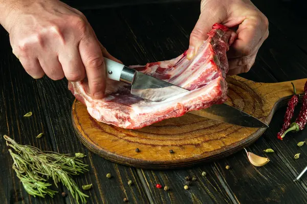 A butcher cuts raw ribs with a knife on a wooden kitchen board. Low key concept of the process of preparing a meat dish for lunch.