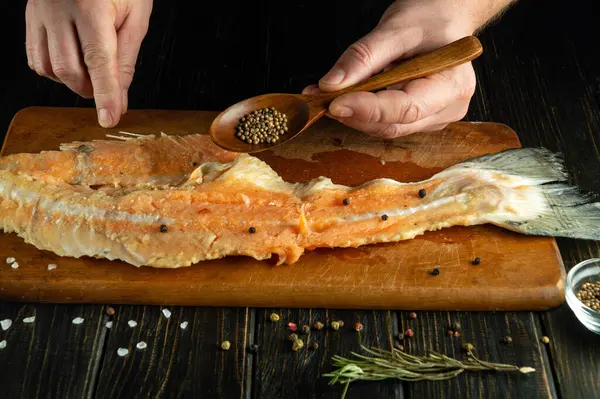 The cook adds coriander with a spoon to the raw fish steak on the kitchen board. Low key concept of preparing fish herring in a restaurant or tavern kitchen.