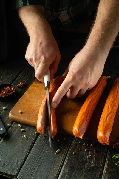 Slicing sausage with a knife in the hand of a cook on a kitchen board before preparing dinner. Low key concept for serving meat dish.