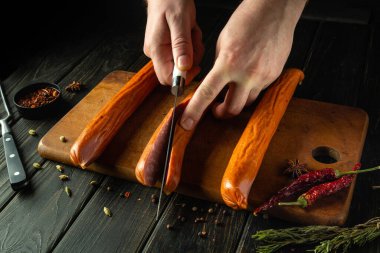 Erkek eller mutfak masasında yemek hazırlamadan önce Bavyera sosislerini bıçakla kesiyor. Et yemeği konsepti. Reklam için karanlık bir yer..