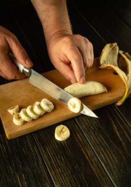 Bir aşçı, mutfak masasında meyve tabağı hazırlamak için olgun bir muzu dilimlemek için bıçak kullanır. Koyu bir arkaplanda reklam için yer.