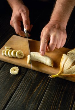 Bıçaklı erkek eller, mutfakta yemek pişirmek ya da işlemek için olgun bir muzu tahta bir tahtada dilimlerler. Kahvaltı için meyve menüsü hazırlama konsepti.