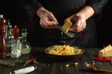 Profesyonel bir aşçı fırçalayıcı kullanarak bir tabak makarnaya sert peynir rendeler. Restoranın mutfağında baharat ve mutfak malzemeleriyle çalışma ortamı.