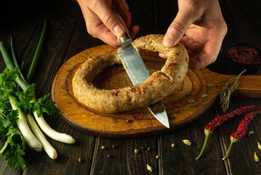 Ahşap kesme tahtasındaki sosisi kesmek için bıçak kullanan bir aşçı elinin yakın çekimi. Otel mutfak masasında ulusal bir et yemeği pişirmek..