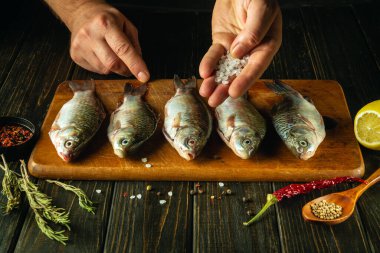 Kesme tahtasındaki çiğ balığa kaba tuz ekleyen şefin yakın çekimi. Mutfak masasında baharat ve kişniş bulunan tuzlu sazan balığı pişirme kavramı..