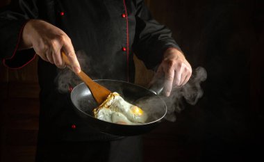 Siyah üniformalı profesyonel bir aşçı elinde kızarmış yumurta ve ahşap bir spatulayla kızartma tavası tutuyor. Çırpılmış yumurta pişirme kavramı. Reklam için yer.