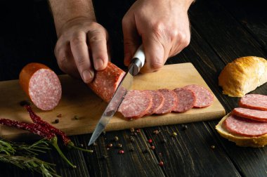 Aşçı elleri, tahta kesme tahtasındaki salam sosisini kesmek için bıçak kullanır. Lokantadaki mutfak masasında akşam yemeği için lezzetli sandviçler hazırlıyorum..