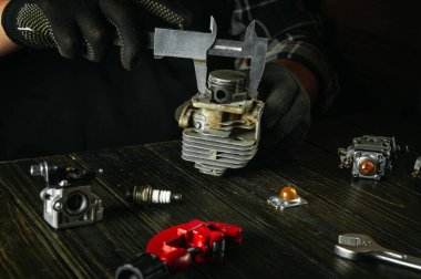 Bir atölyedeki çalışma masasına yakın çekim yapan bir zanaatkârın elleri eski bir çim biçme makinesinin veya budayıcının motorunu tamir ediyor. Piston çapını kalibre ile ölçmek.