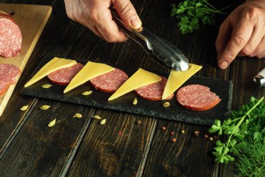 Şefin elleri siyah mutfak tahtasında servis yaparken sosisin üzerine maşa koyuyor..