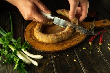 Ahşap bir kesme tahtasındaki et sosisini kesmek için bıçak kullanan bir şefin yakın çekimi. Mutfak masasında ulusal bir et yemeği pişirmek..