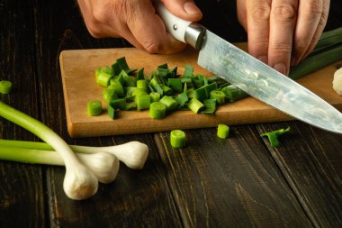Sarımsaklı vejetaryen yemeği hazırlıyorum. Şefin elleri, tahta kesme tahtasındaki genç yeşil sarımsağı kesmek için bıçak kullanıyor. Reklam için yer.