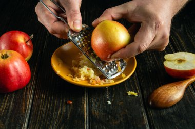 Bir aşçı, ellerindeki el rendesiyle taze bir elmayı elmalı tatlı yapmak için mutfak masasında rendeliyor..