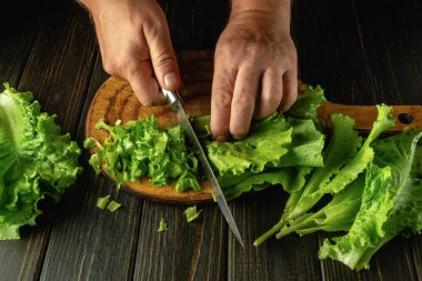 Marul yapraklarını kesmek için kesme tahtasındaki bıçağı kullanan erkek eller evde öğle yemeği için sebze tabağı hazırlıyor..