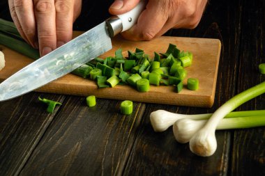 Bir aşçı, akşam yemeğini hazırlamadan önce ahşap bir mutfak tahtasındaki yeşil sarımsağı kesmek için bıçak kullanır..
