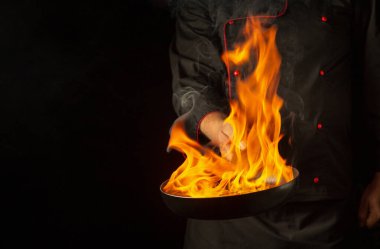 Kızartma tavasında ateş ya da alevle yemek pişiren bir aşçı. Barda öğle yemeği pişirme kavramı. Siyah arkaplanda tarif için boşluk.