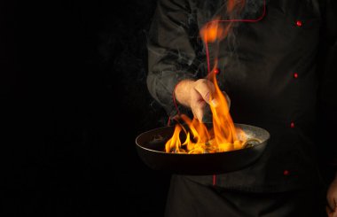 Aşçı alevlerle yanan bir tavada sebze pişiriyor. Siyah arka planda restoran ve otel servisi kavramı. Flambe ya da doğal ateşle pişirme.
